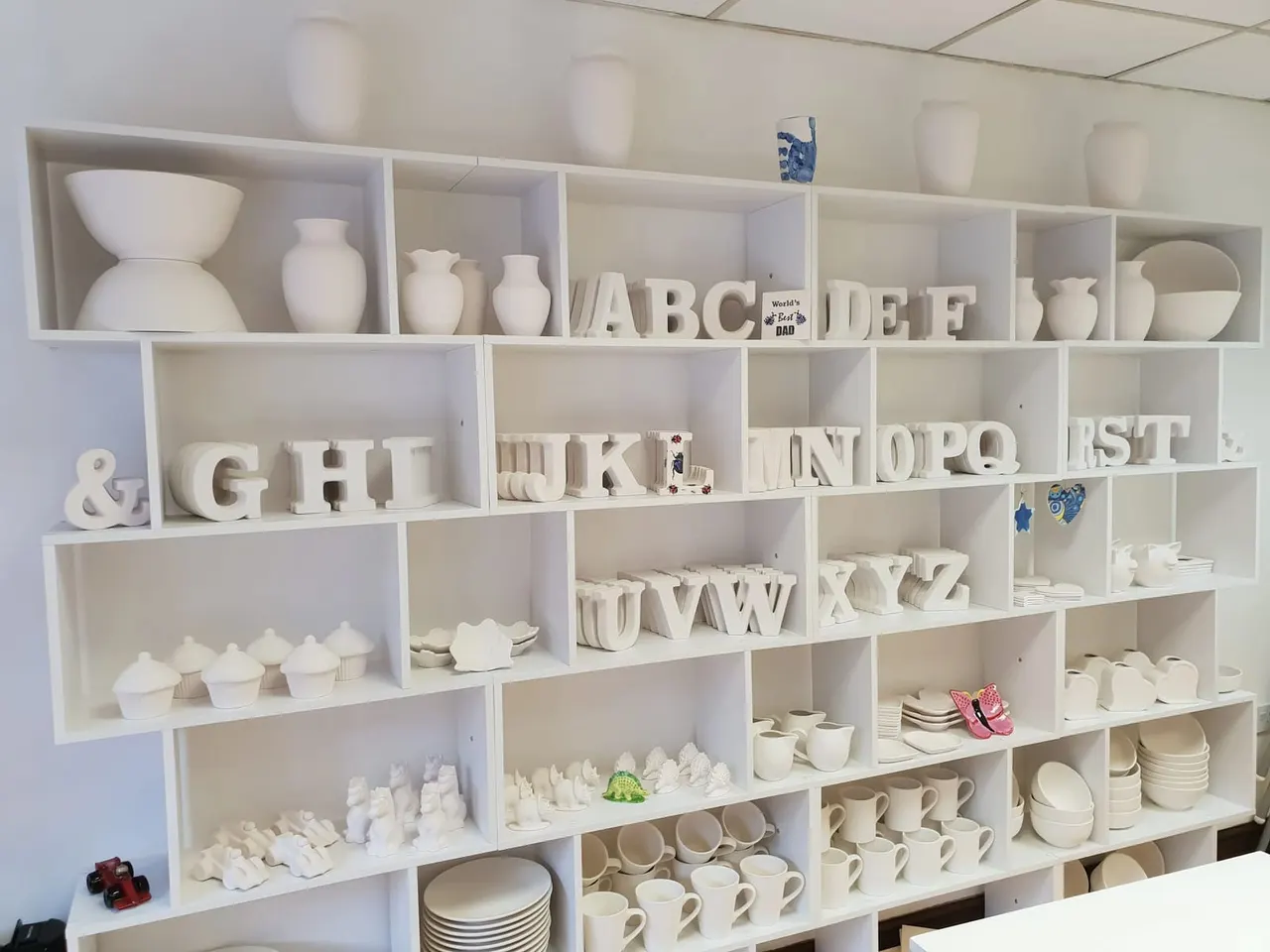 White shelving unit displaying ceramic tableware, decorative vessels, alphabet letters, and small decorative items.
