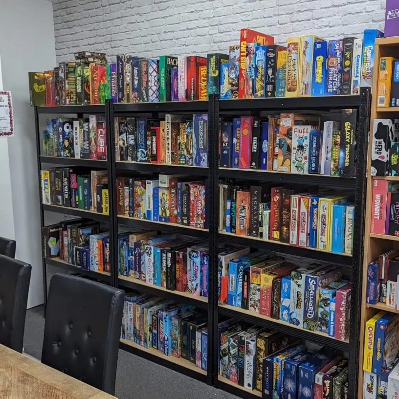 Black shelving units displaying a large collection of colourful boxed board games arranged on multiple shelves against a white brick wall.