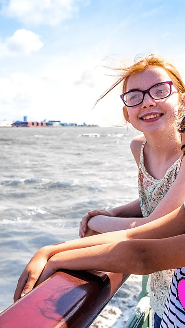 Young child wearing glasses smiling at the sea with a fairground visible on the horizon.