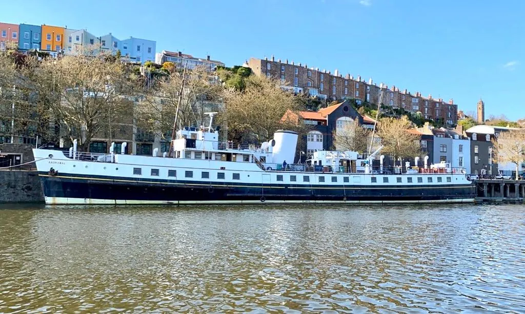 A vintage passenger ship moored on a river beside colourful waterfront buildings on a hillside.