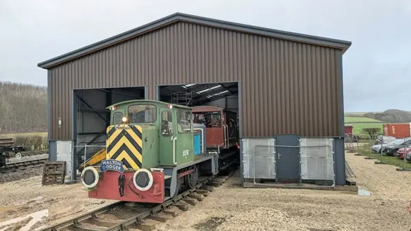 Small heritage railway locomotive emerging from corrugated iron shed in rural countryside setting.