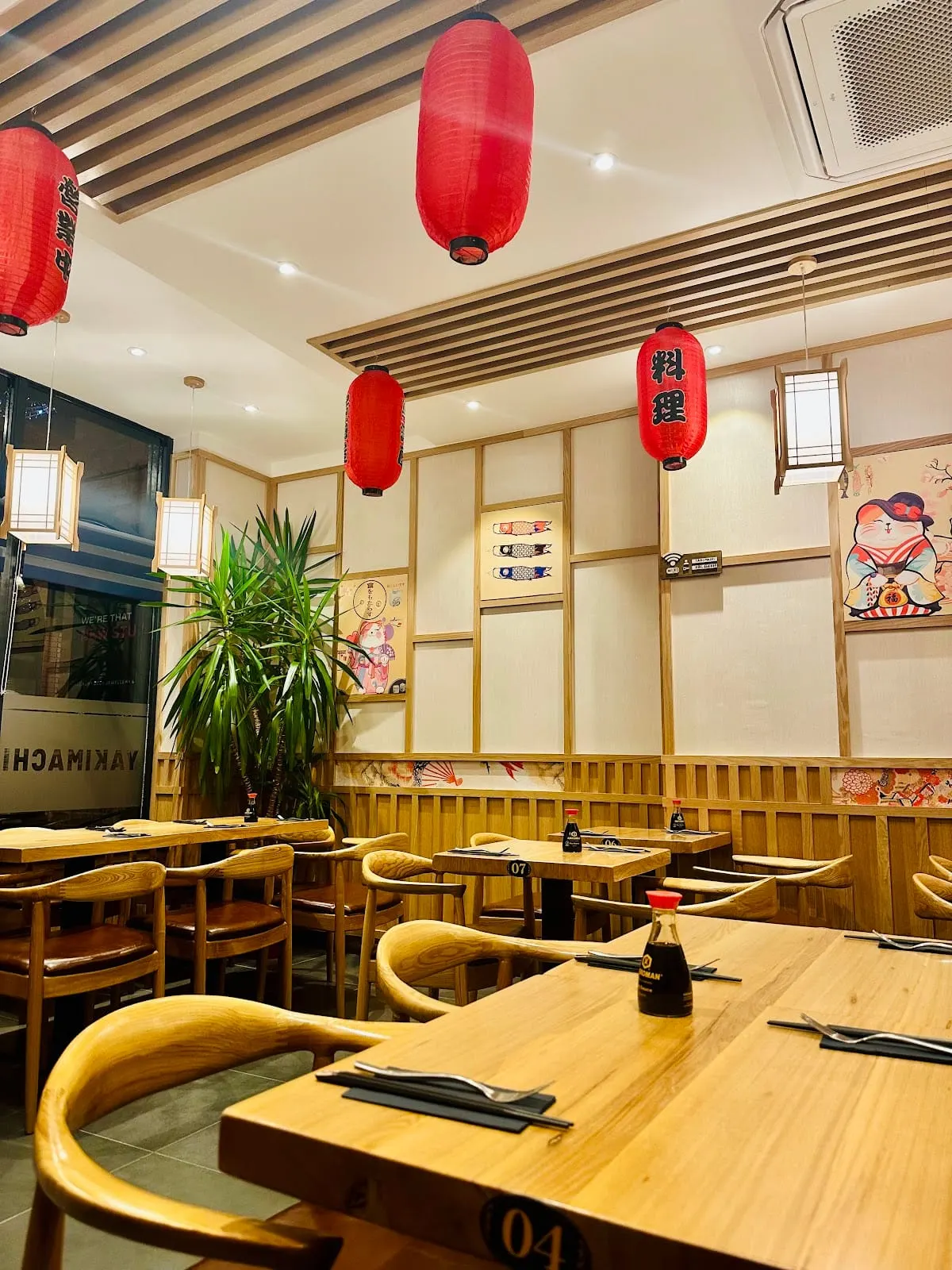 Interior of a Japanese restaurant with wooden tables, chairs, and panelling, decorated with red paper lanterns and traditional artwork.