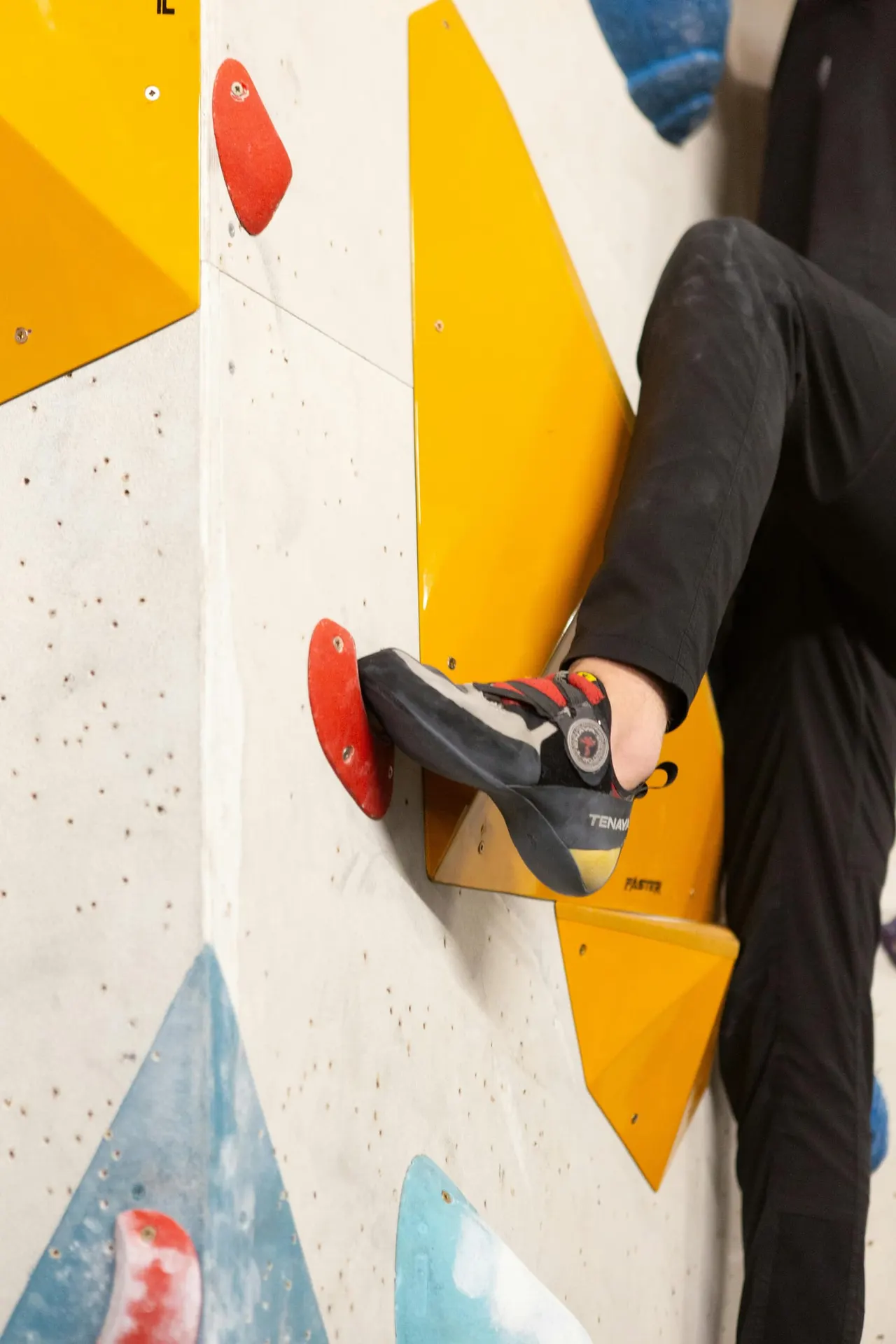 Child climbing an indoor wall with red and yellow hand holds whilst wearing climbing shoes and safety equipment.