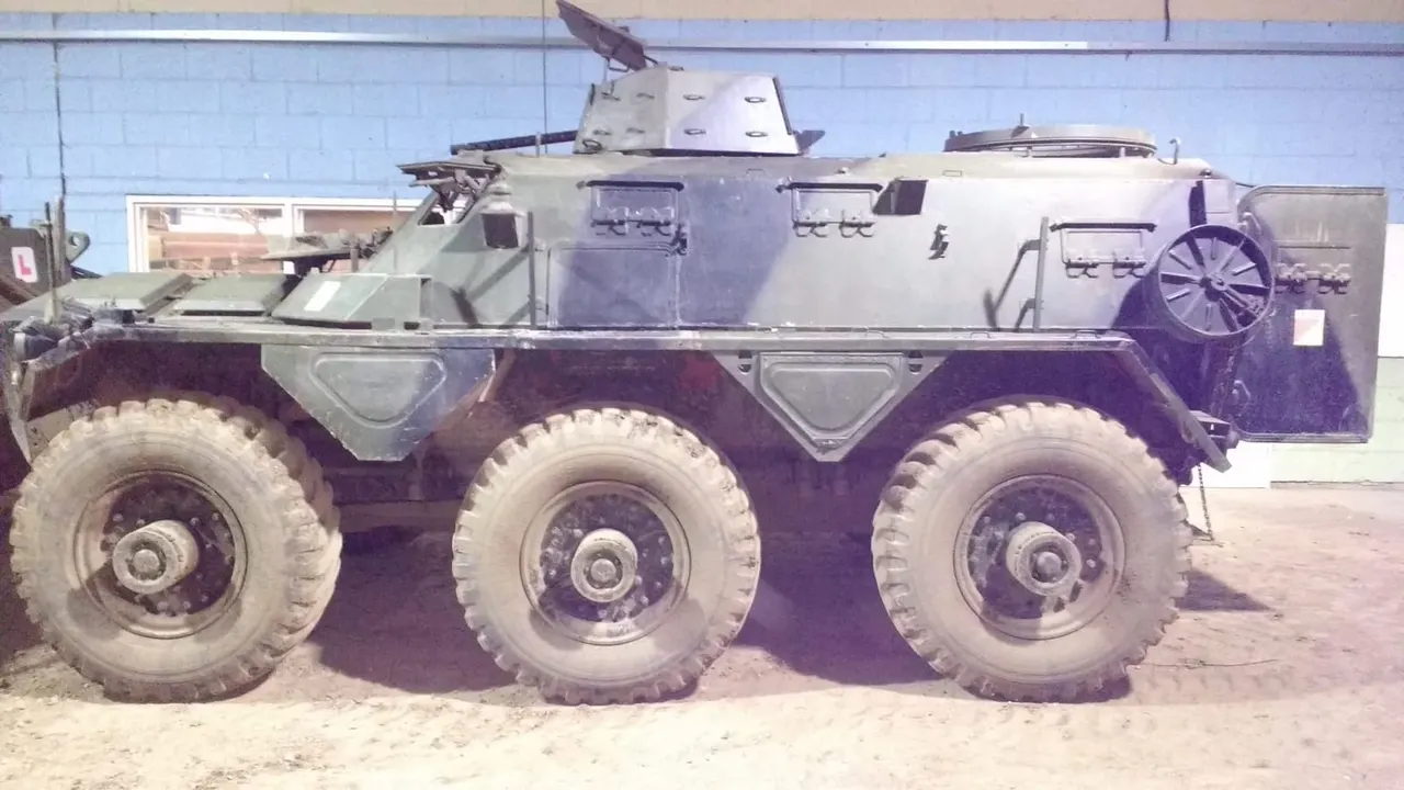 Military armoured personnel carrier with six large wheels displayed outdoors in a sandy setting.