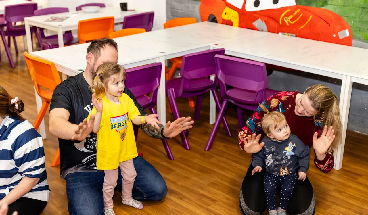Adults and toddlers playing together in a colourful indoor soft play facility with purple and orange chairs and play equipment.