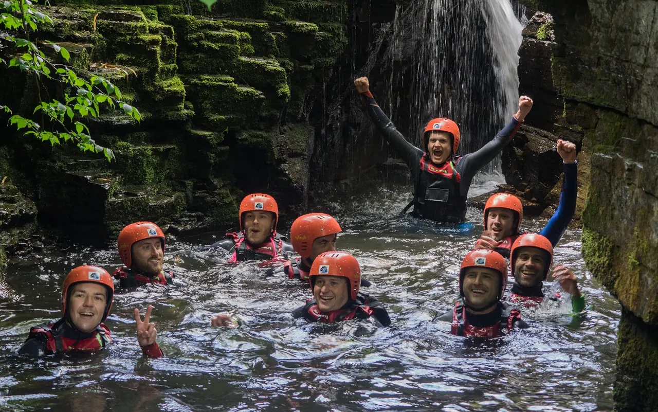 Group of people wearing orange helmets and wetsuits in a gorge stream beneath a waterfall.