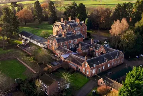 Aerial view of a large brick mansion with surrounding grounds, trees, and manicured lawns in countryside setting.