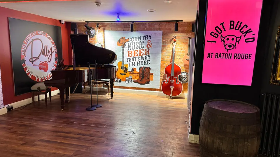 An empty country music venue with a grand piano, acoustic guitars, and a cello on stage, with neon signage and a wooden barrel in the foreground.
