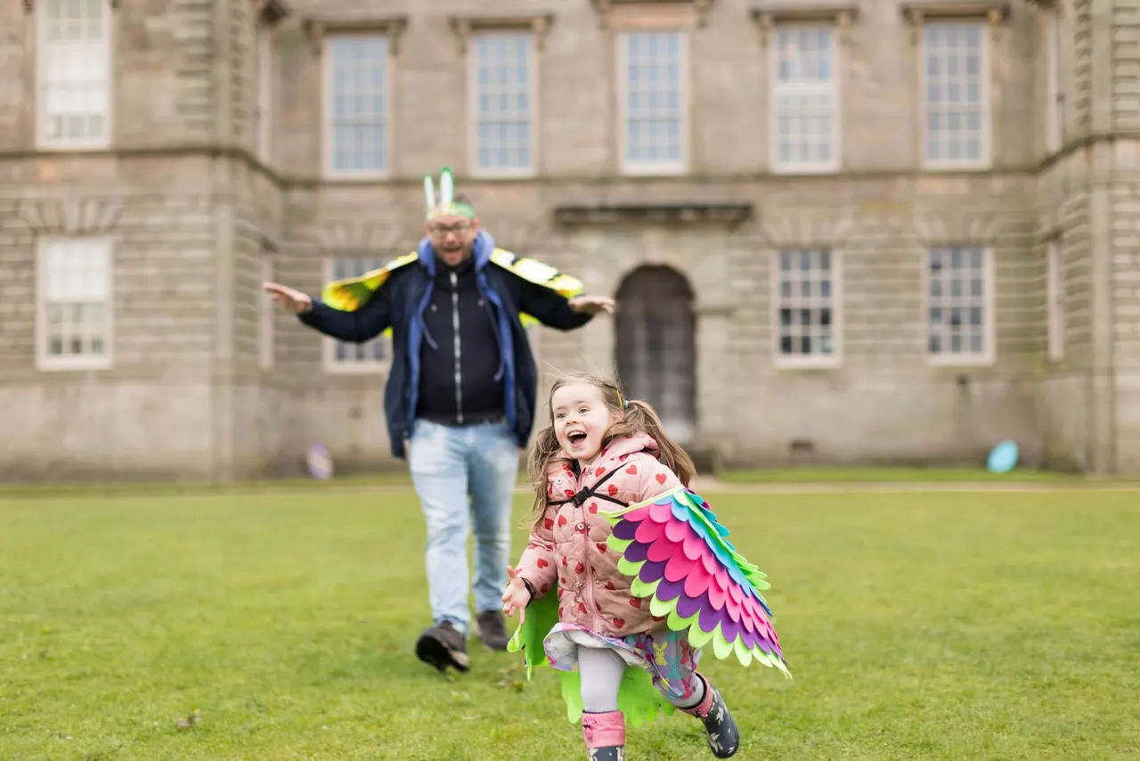 Adult and child in fancy dress running across a lawn in front of a historic stone building.