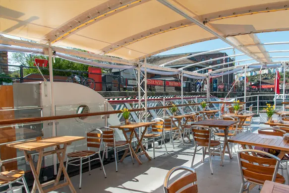 Outdoor dining area on a covered deck with wooden tables and chairs, overlooking a harbour with boats and waterfront buildings.
