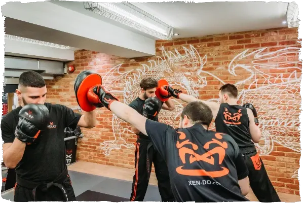 Multiple martial artists in black and orange gear practising kick boxing drills against a brick wall.