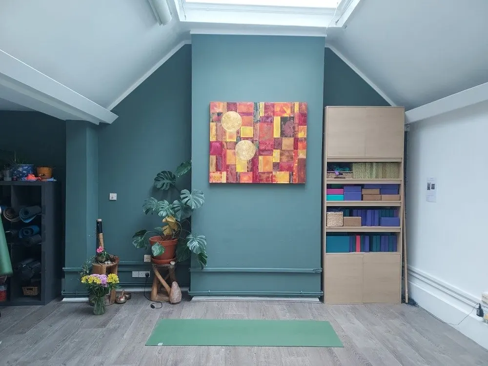 A bright, modern wellness studio with teal walls, wooden flooring, a green yoga mat, potted plants, and colourful artwork, ready for classes.
