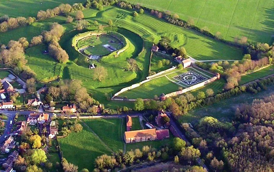 Aerial view of historic estate ruins with circular earthwork fortifications, formal gardens and surrounding parkland with trees.