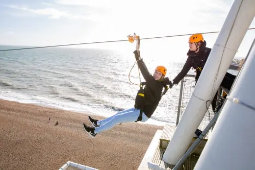 Person in safety gear ziplining across a beach towards the shore, with instructor watching from platform.