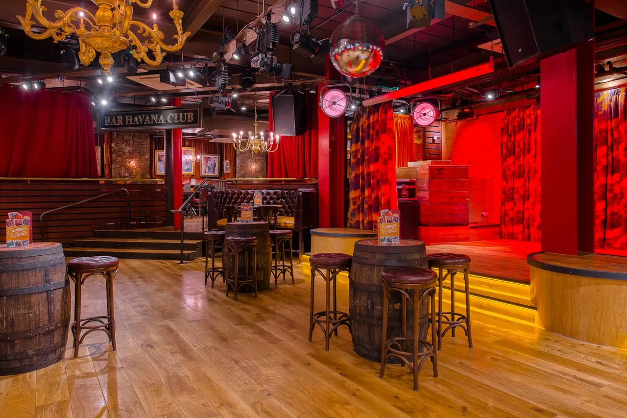 Interior of a nightclub with red lighting, barrel-style tables, bar stools, a stage area, and a "Bar Havana Club" sign.