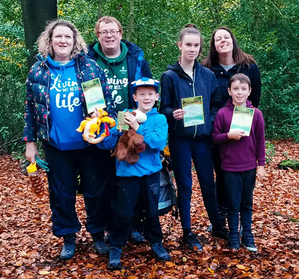 A family group of six people and a small dog posing outdoors holding activity cards against a leafy green hedge backdrop.