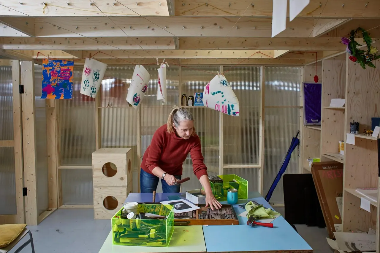 Woman working at a crafts table in a wooden studio space with hanging artwork and storage.