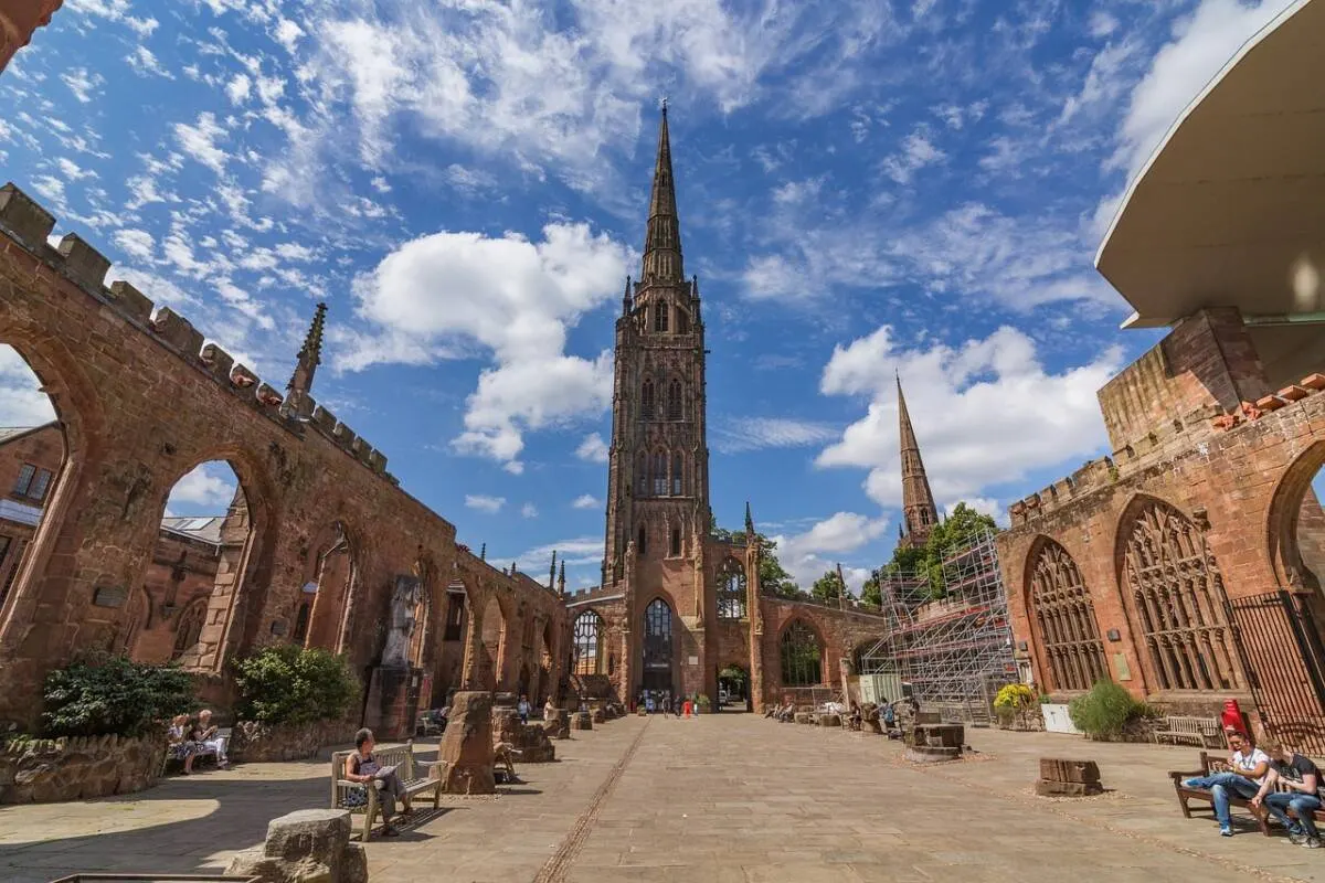 Red sandstone ruins of a medieval church with a tall spire, surrounded by visitor seating in a sunny courtyard.