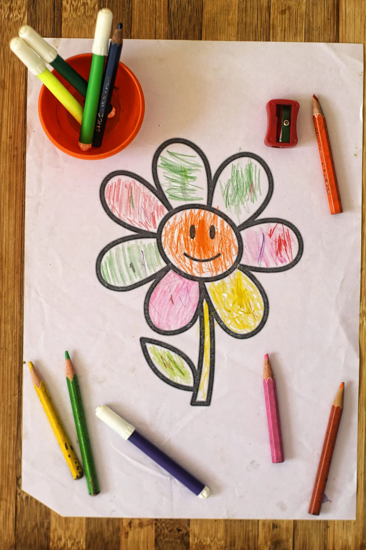 Child's coloured pencil drawing of a smiling flower with multi-coloured petals on white paper, surrounded by pencils and a sharpener on wooden surface.