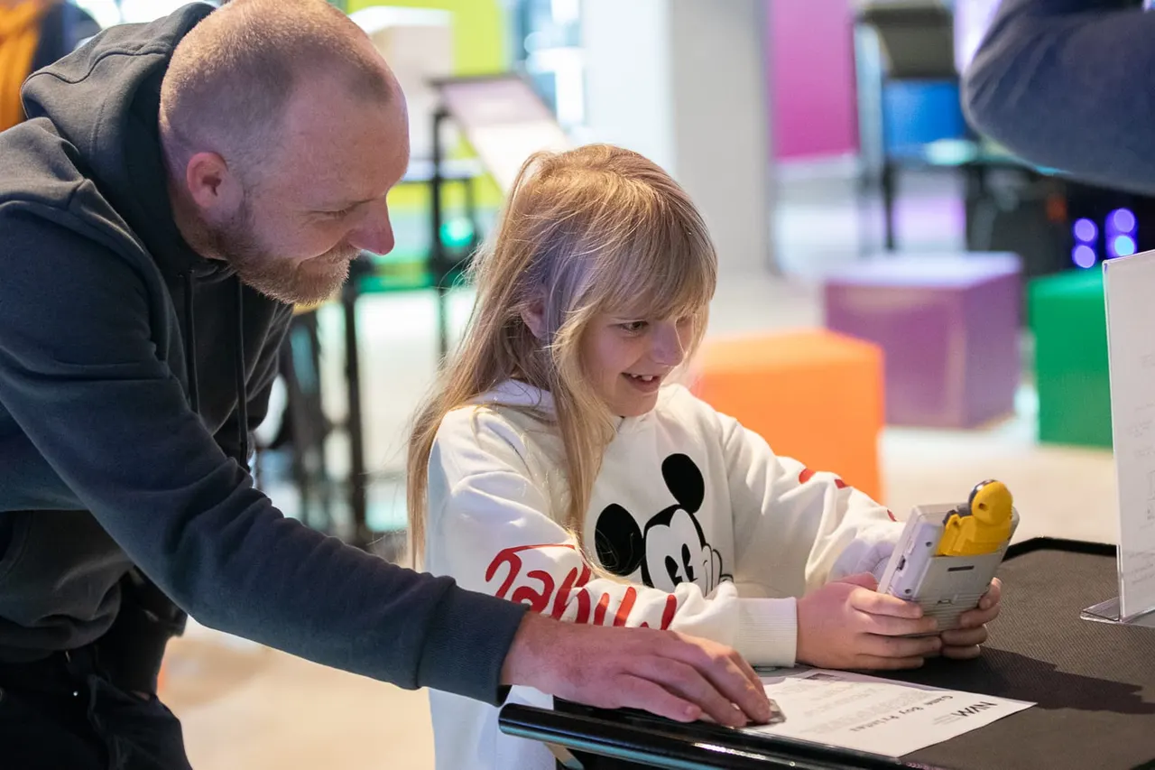 Adult and child interact with a touchscreen display in a colourful indoor museum space with game-related exhibits.