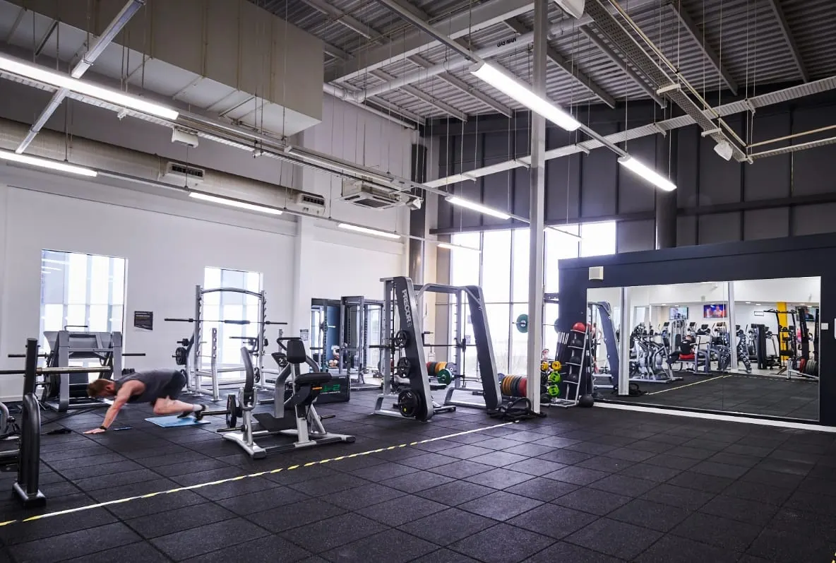 Person exercising on mat in modern gym with weight machines, mirrors and fluorescent lighting throughout.