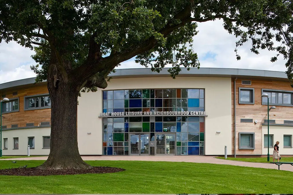Modern leisure centre building with wooden cladding and large coloured windows beneath a mature tree on a grass lawn.