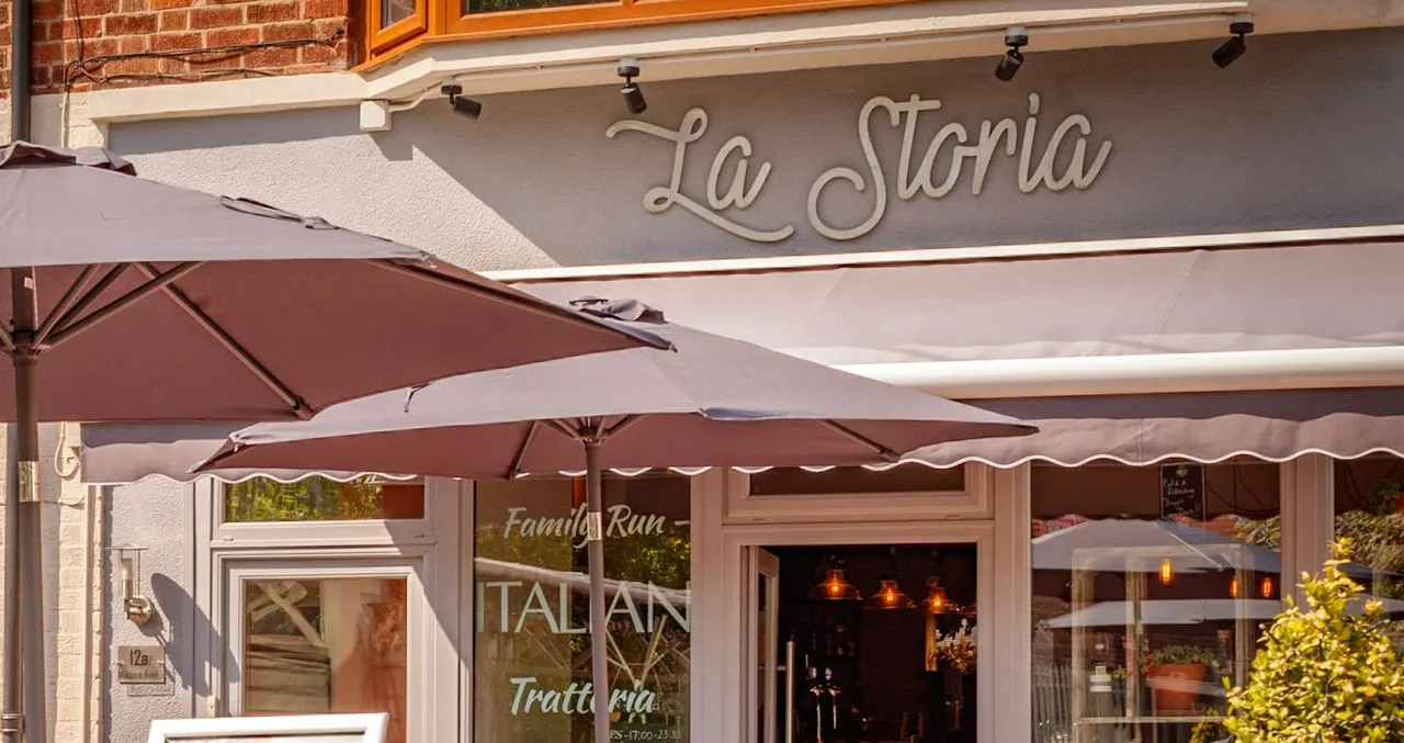 Exterior storefront of La Storia restaurant with burgundy umbrellas, signage, and warm lighting visible through windows.