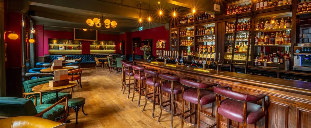 Interior of an upscale bar and lounge with wooden bar counter, burgundy leather stools, colourful seating booths, and extensive spirit collection behind the bar.