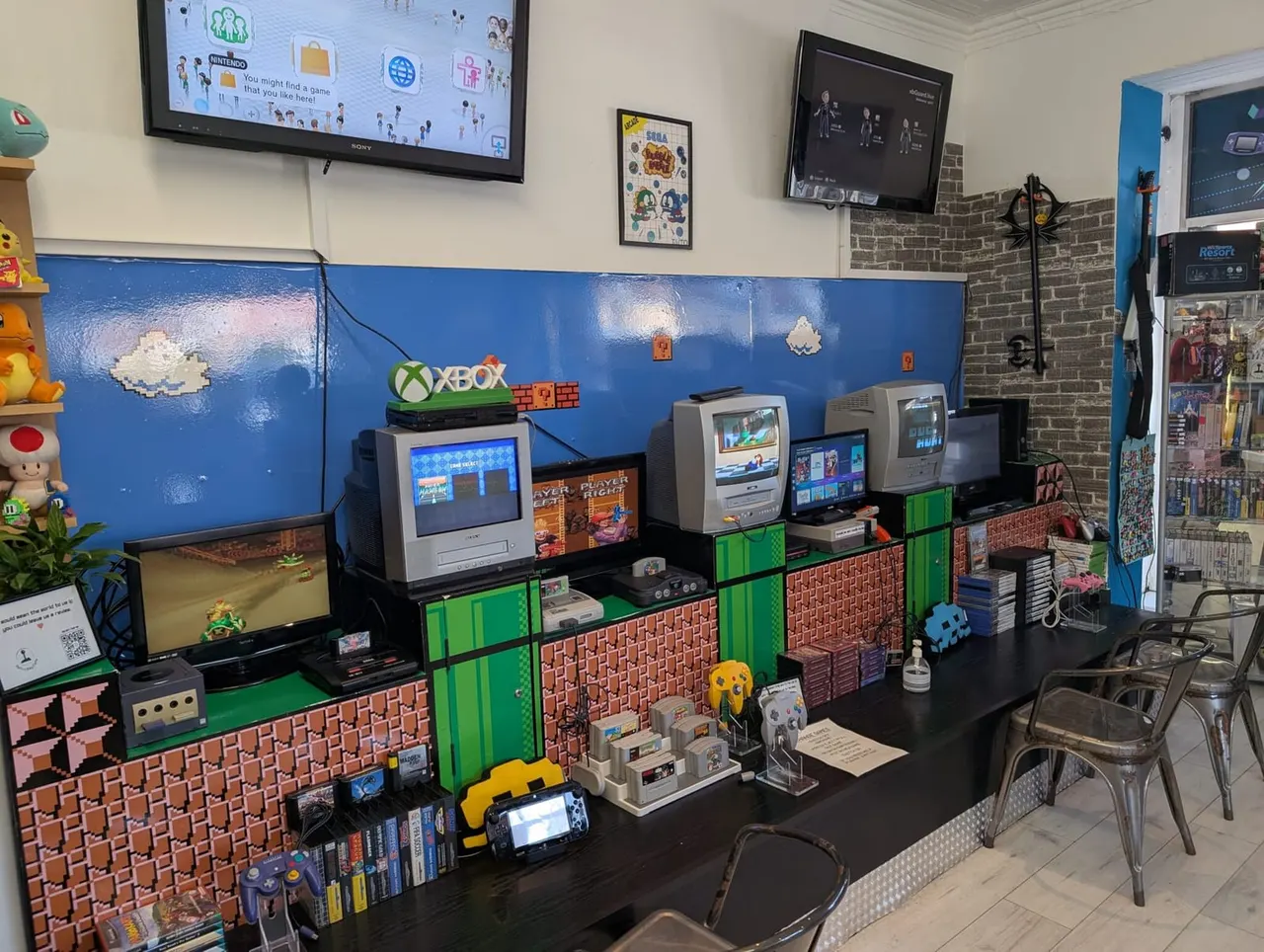 A retro gaming café interior with multiple vintage gaming consoles and screens displayed on shelving units decorated with green and brick-patterned panels.