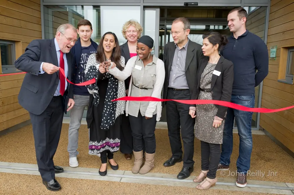 A group of eight people cutting a red ribbon at the entrance of a modern building during an official opening ceremony.