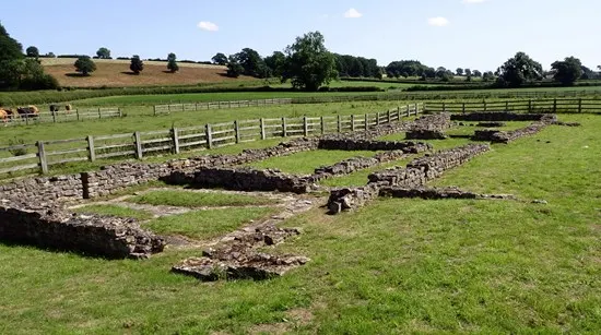 Photograph

Excavated Roman foundations and stone outlines marked by rocks on a grassy field with wooden fencing.