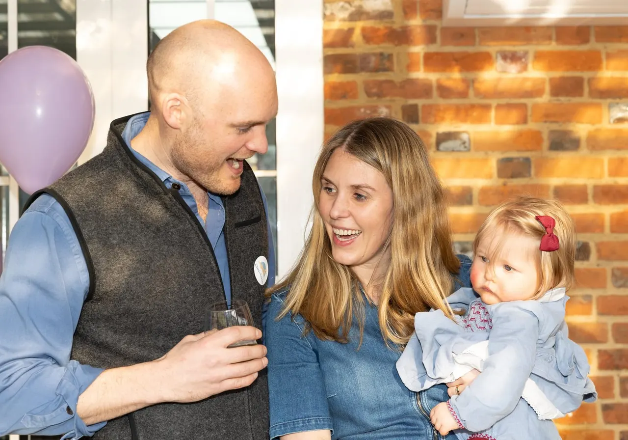 A man and woman with a young child laugh together at an indoor celebration with a purple balloon and brick wall backdrop.