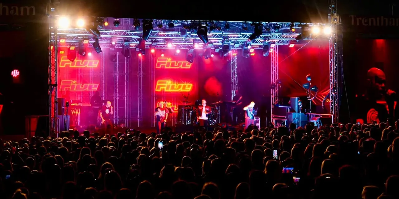 A large crowd watches a live band performing on a brightly lit stage with red and purple neon signage at night.