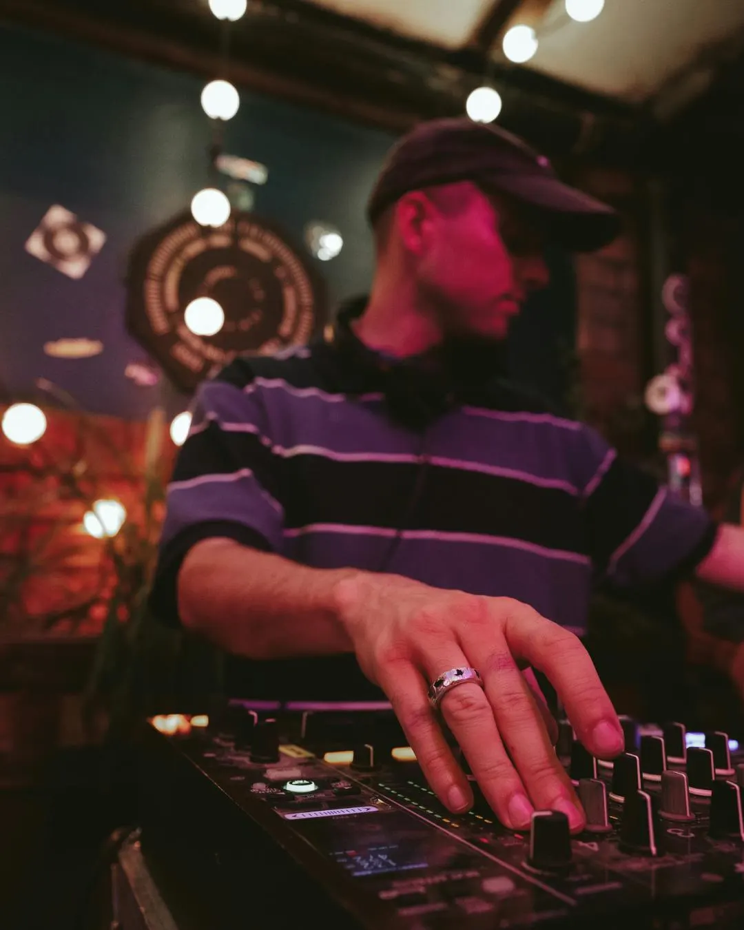 DJ operating decks at a nightclub with purple and red lighting and overhead bulbs.