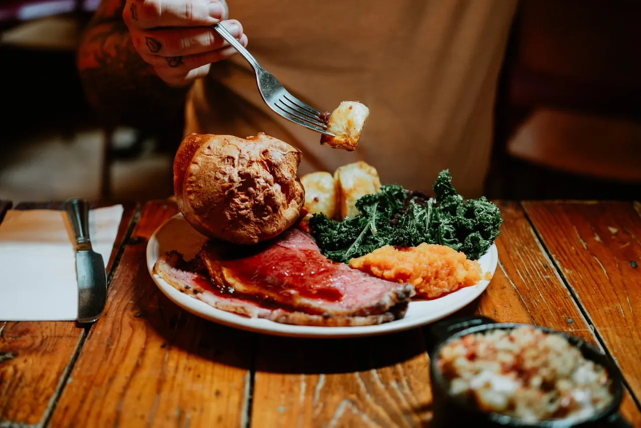Person in red tattoo sleeve holding fork above plate of sliced meat, roasted vegetables and kale at wooden table.