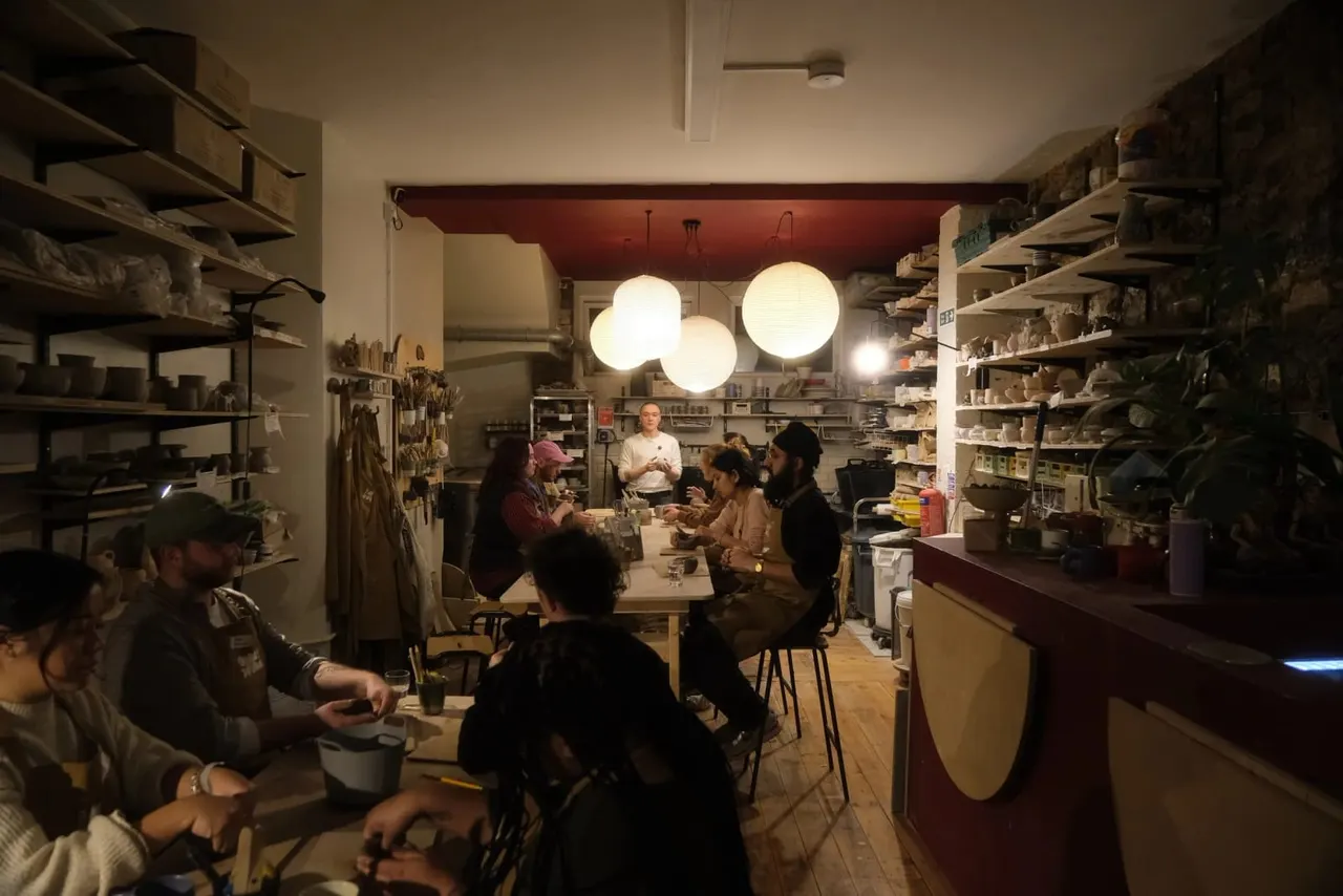 People seated at tables in a pottery studio workshop, surrounded by shelves of clay materials and art supplies, with large pendant lights overhead.