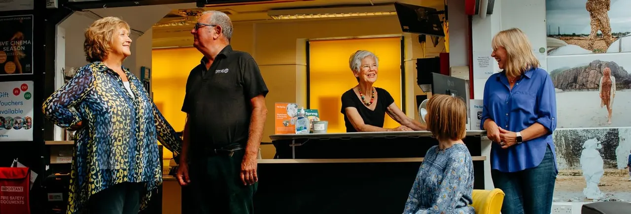 Staff member at reception desk assists visitors, including a child, in the leisure centre foyer.