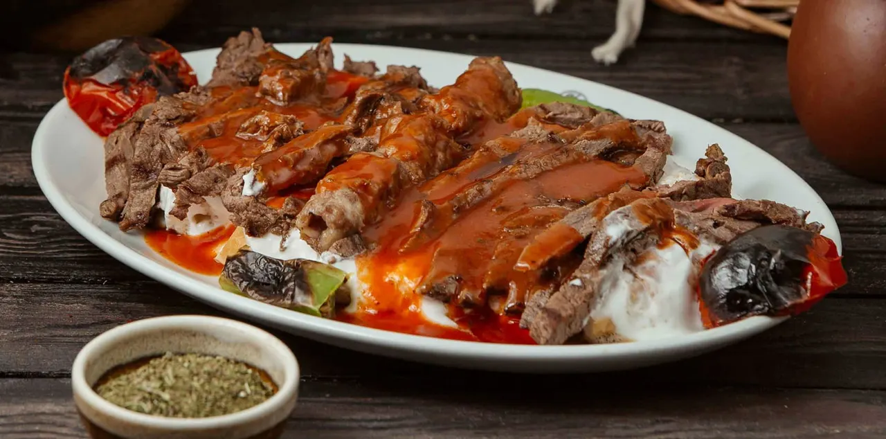 Sliced grilled meat with peppers, tomatoes and yogurt on a white plate, served with a small bowl of sauce.