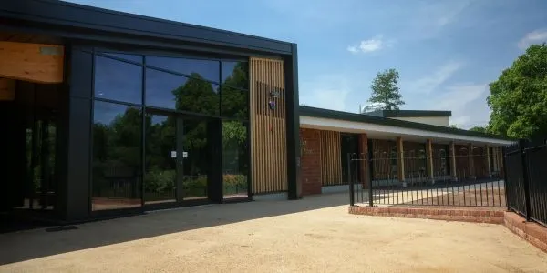 Modern single-storey building with dark frame and large windows overlooking a sandy courtyard and green surroundings.