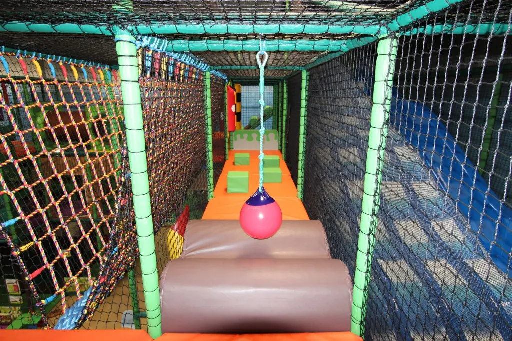 Indoor soft play tunnel with netting sides, foam floor, and a suspended pink ball toy in an enclosed play area.