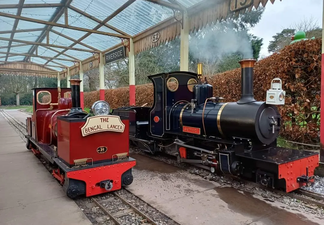 Two miniature steam locomotives on narrow-gauge tracks under a glass canopy, with hedging and signage visible in the background.
