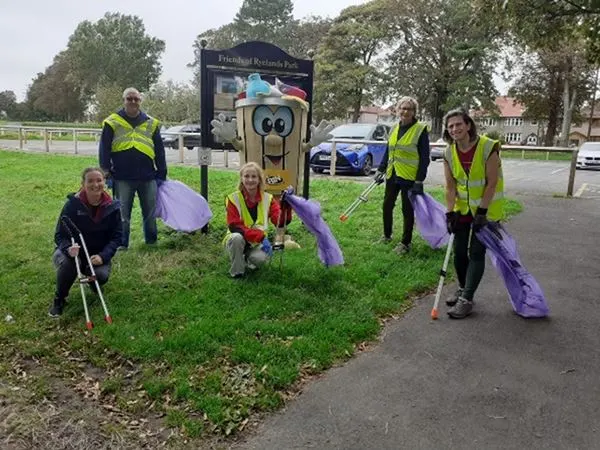 A group of volunteers in high-visibility clothing collecting litter at a park, holding purple bags near an information sign.