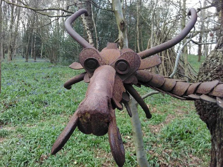 Rusted metal sculpture of a creature with horns and outstretched limbs amongst woodland trees and undergrowth.