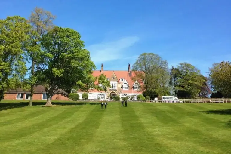 A large brick manor house with terracotta roof surrounded by open parkland, mature trees and white fencing, with a few people visible on the expansive lawn under clear blue sky.