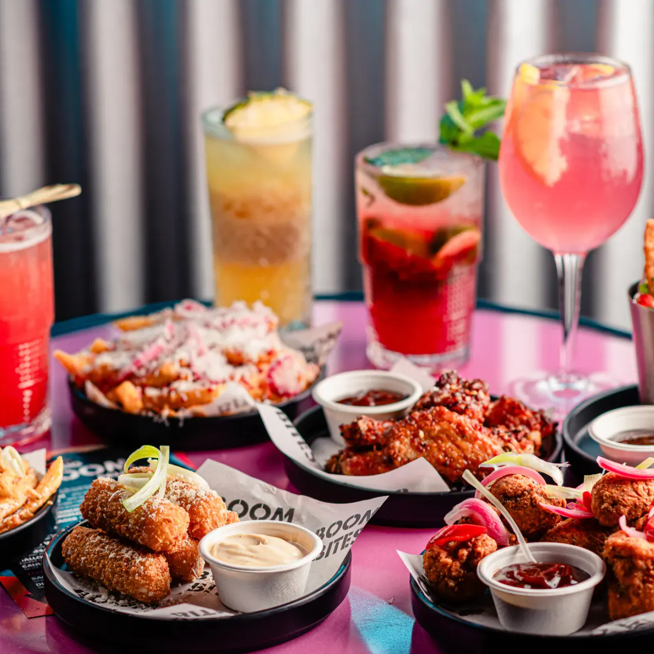 Colourful cocktails and fried food platters arranged on a pink table at a bar venue.