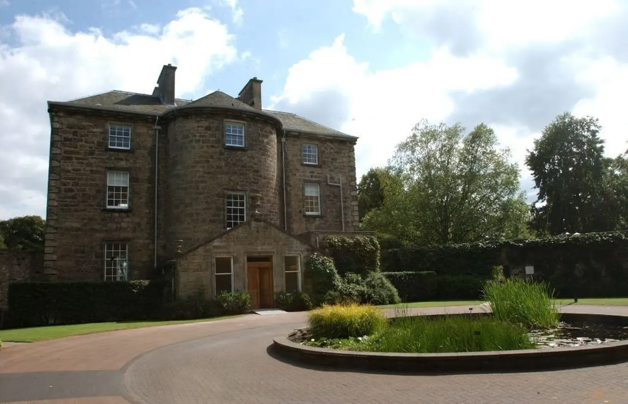 Stone mansion with curved driveway, central planted garden bed, and surrounding hedges under blue sky.