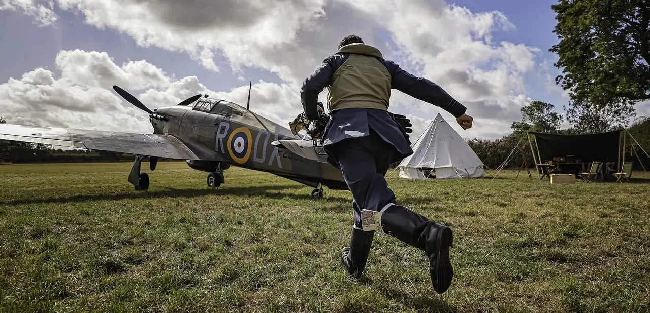 Person in period military uniform running towards a vintage RAF aircraft on a grass airfield with canvas tent and trees visible.