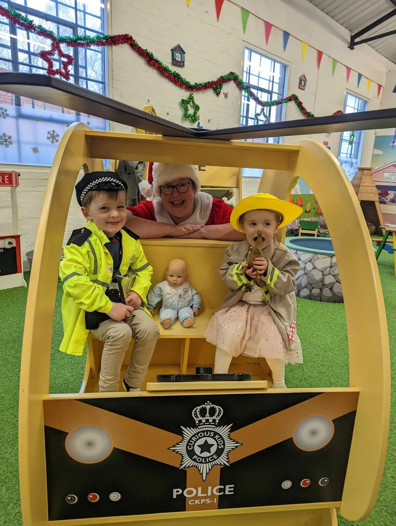 An adult and two young children in fancy dress sit inside a large yellow toy police van with a doll, in an indoor play centre decorated with festive garlands.