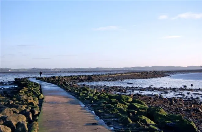 Person walking along moss-covered rock breakwater extending into calm coastal waters with distant hills.