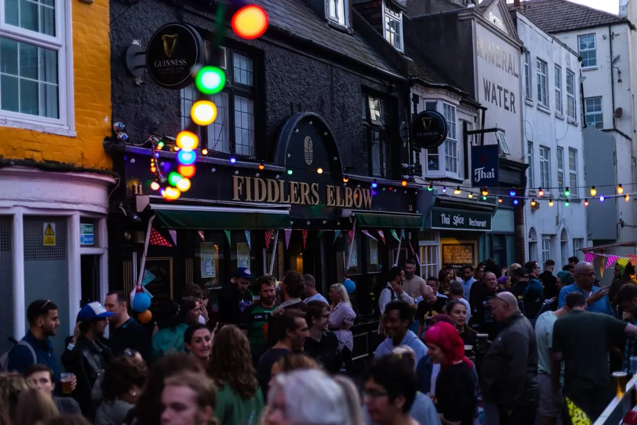 Crowded street scene outside Fiddlers Elbow pub with colourful festive lights and bunting, groups of people socialising outside the venue.